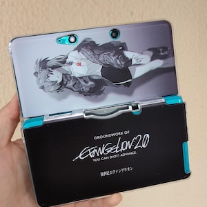 Può includere: Custodia per console di gioco portatile nera e verde acqua con grafica anime. La custodia presenta il testo "GROUNDWORK OF EVANGELION 2.0" e testo giapponese. La console è aperta, rivelando lo schermo con il personaggio anime.