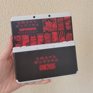 Può includere: Una console di gioco portatile bianca e nera con un tema One Piece. L'area dello schermo superiore mostra il testo SUPERNOVA e illustrazioni di personaggi anime in rosso. La sezione inferiore presenta il logo One Piece in rosso con icone.