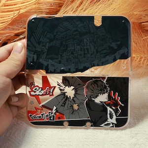 May include: Transparent plastic Nintendo 3DS case. The top half has a black and white cityscape design. The bottom half features a character graphic with red accents, and the words "Show Time" and "Slot!" in red and white.