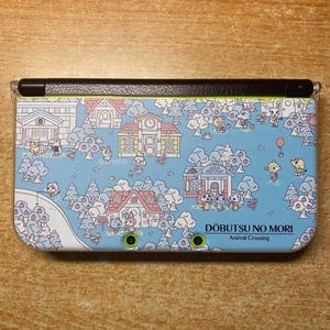 Op de afbeelding: Nintendo 3DS handheld gameconsole met een lichtblauwe hoes. De hoes heeft een kleurrijke illustratie van een dorpsscène met cartoon dieren, bomen en gebouwen. De tekst "Dobutsu No Mori Animal Crossing" is zichtbaar.