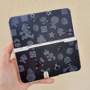 May include: A black Nintendo 3DS handheld game console with a clear protective case. The console is decorated with gray cartoon characters and icons from the Super Mario Bros. video game series.