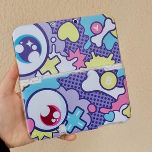 Puede incluir: Una funda de consola de juegos portátil estampada. La funda presenta un diseño vibrante con ojos de dibujos animados, corazones y formas de huesos en tonos morado, azul, rosa y amarillo. La funda está abierta, revelando el interior.