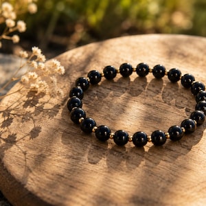 May include: A circular beaded bracelet featuring smooth, polished black spherical beads alternating with small, reflective gold-toned metallic spacer beads. The bracelet is arranged in a continuous loop, showcasing a uniform design of dark stones.