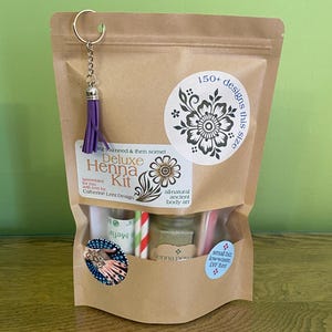 May include: A brown paper bag with a clear plastic window, containing a henna kit. The kit includes henna powder, a small bottle of oil, two straws, a small bottle of mefix, and a small blue and white sticker that says "small biz lowwaste DIY fun!" The bag has a purple tassel attached to a silver key ring.