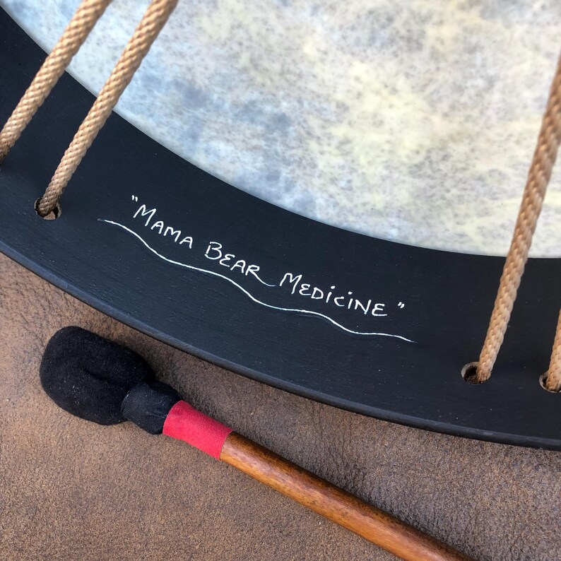 May include: A round frame drum with a light-colored, textured drumhead and a black wooden frame. The frame features the white hand-painted text "MAMA BEAR MEDICINE" and is laced with tan rope. A wooden mallet with a black head and red tape is included.