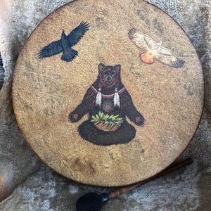May include: A round, tan-colored drum featuring a central illustration of a brown bear sitting with a basket of greenery, flanked by a black raven on the left and a hawk on the right. A wooden mallet with a black head rests below the drum.