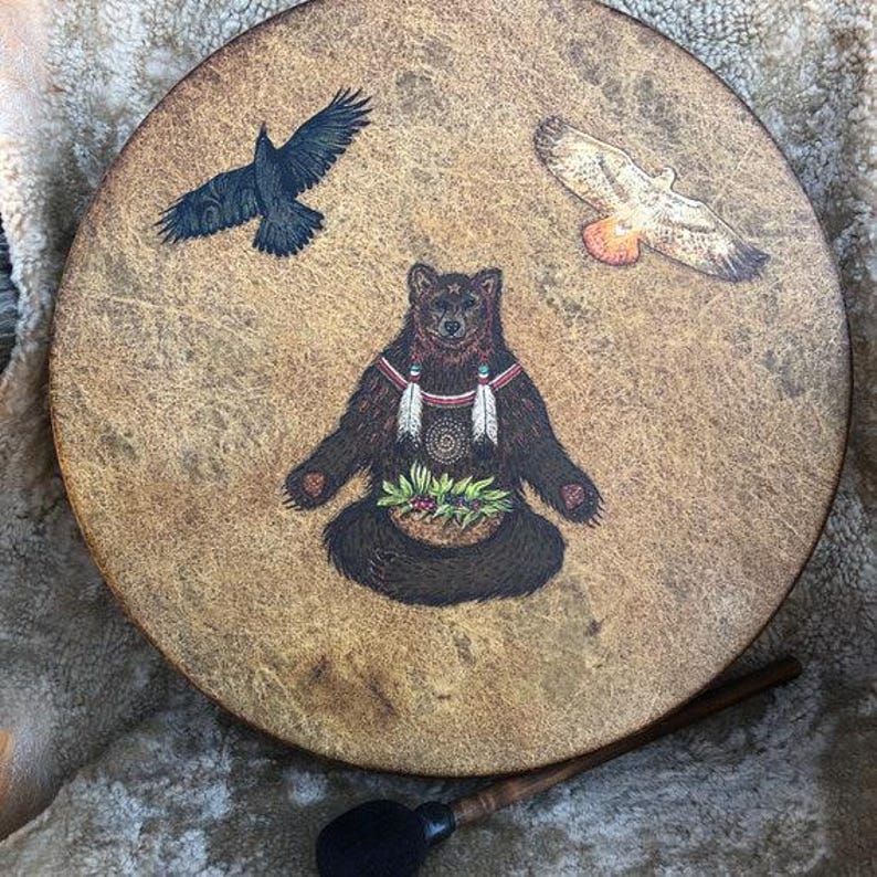 May include: A round, tan-colored drum featuring a central illustration of a brown bear sitting with a basket of greenery, flanked by a black raven on the left and a hawk on the right. A wooden mallet with a black head rests below the drum.