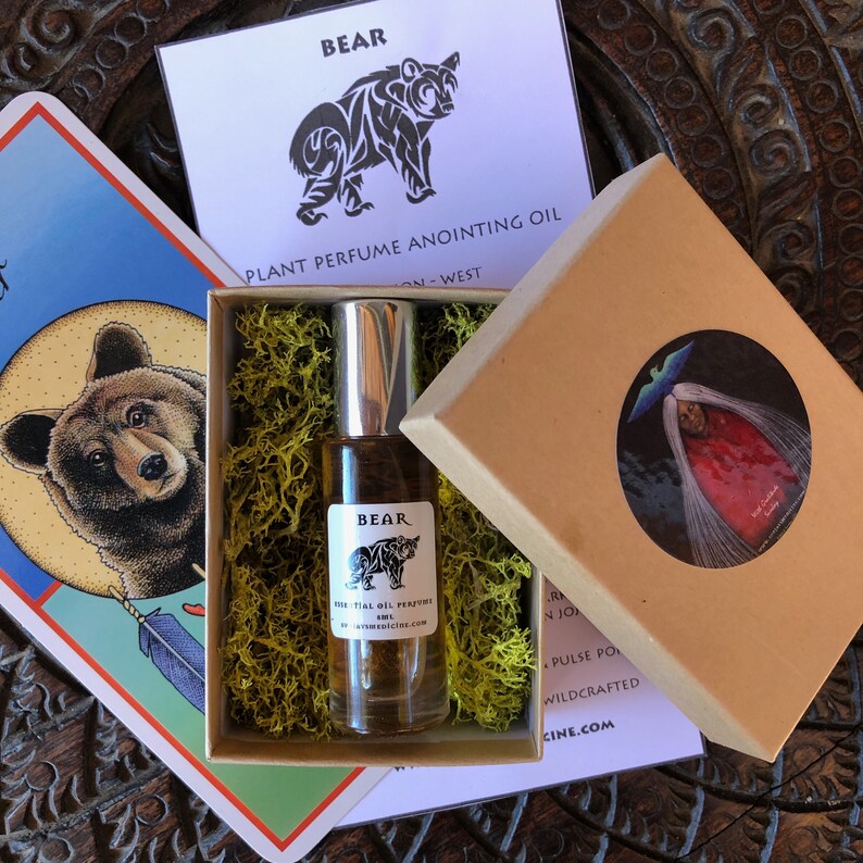 May include: A glass roller bottle of Bear plant perfume anointing oil with a silver cap, nestled in a cardboard box filled with green moss. The label features a black bear illustration and text. The box lid displays a circular sticker with a figure.