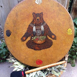 May include: A round drum with a tan, textured surface featuring a central illustration of a brown bear holding a basket of greenery. The bear is adorned with feathers and a spiral design. Four colored bear paw prints surround the central image.
