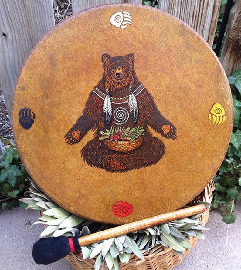 May include: A round drum with a tan, textured surface featuring a central illustration of a brown bear holding a basket of greenery. The bear is adorned with feathers and a spiral design. Four colored bear paw prints surround the central image.