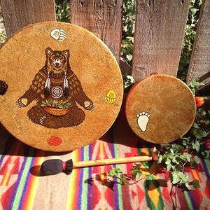 May include: Two round tan drums with rawhide heads and a wooden mallet with a black fabric head. The larger drum features a brown bear illustration with a basket, feathers, and four colored paw prints. The smaller drum has one white paw print.