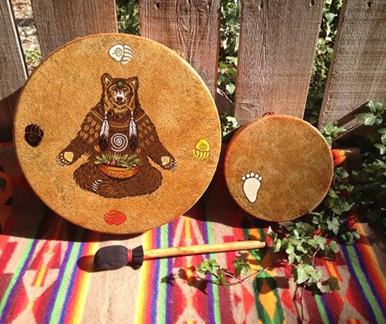 May include: Two round tan drums with rawhide heads and a wooden mallet with a black fabric head. The larger drum features a brown bear illustration with a basket, feathers, and four colored paw prints. The smaller drum has one white paw print.