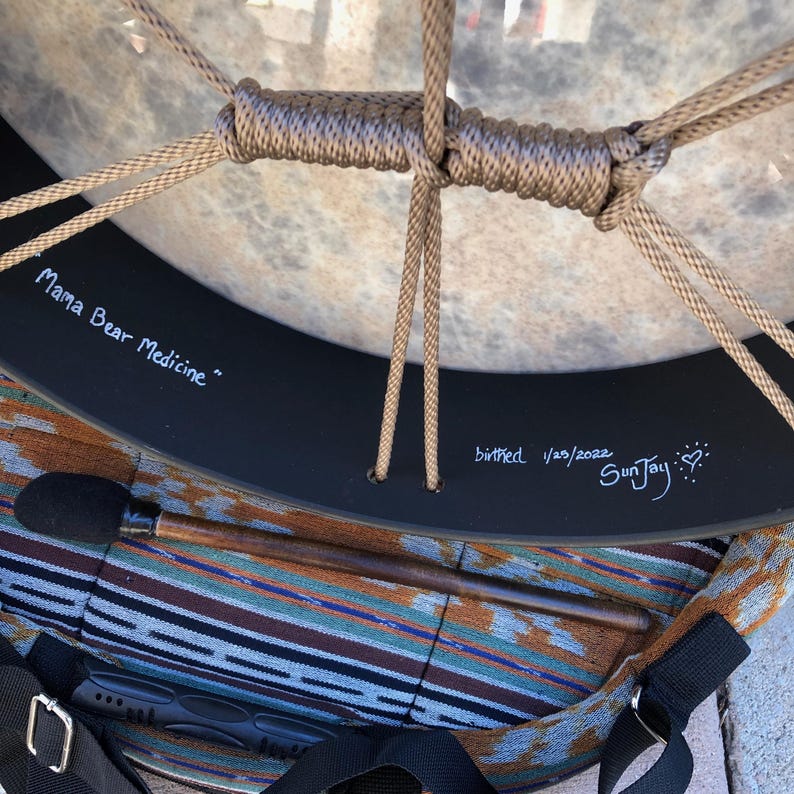 May include: A round frame drum with a light-colored drumhead and tan cord lacing. The black frame features white text reading Mama Bear Medicine, birthed 1/25/2022, and SunJay with a heart symbol. A wooden mallet with a black head rests below.
