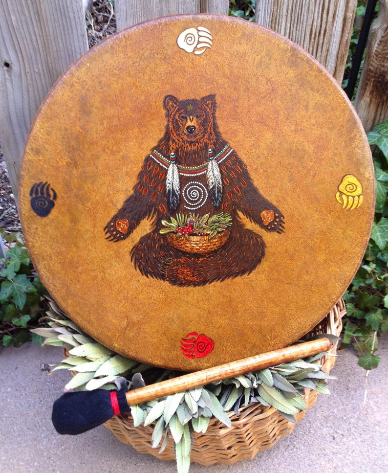 May include: A round drum with a textured tan surface featuring a central illustration of a brown bear holding a basket of berries and herbs. Four colored bear paw prints with spiral centers are positioned at the top, bottom, left, and right.