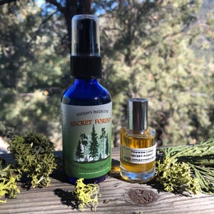 May include: Two glass bottles of plant medicine spray and perfume. The larger blue bottle is labeled "Secret Forest" with a forest graphic. The smaller bottle is labeled "Powwow Lodge Secret Forest". The bottles sit on a wooden surface with greenery.