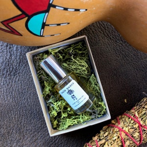 May include: A small bottle of "Rain Dance" palm perfume oil in a white box with green moss. The bottle has a silver cap and a white label with a black and white illustration of a person dancing in the rain.