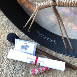 May include: A round drum with a black frame and tan rope lacing, featuring the text Mama Healing Bear. Included are a wooden mallet with a black head, a small packet labeled Bear Plant Perfume Anointing Oil, a rolled paper, and a sage bundle.