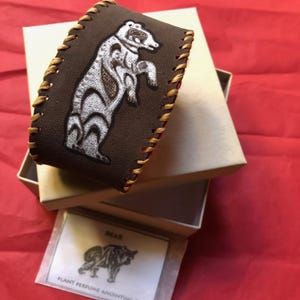 May include: A brown leather cuff bracelet featuring a silver-colored bear graphic and tan whip-stitched edges. Included is a small white card with a bear illustration and the text BEAR PLANT PERFUME ANOINTING OIL printed in black ink.