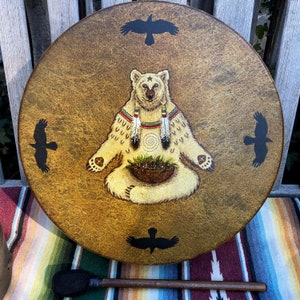 May include: A round, tan-colored drum featuring a central illustration of a seated bear with a star on its forehead, wearing feathers and holding a basket of berries. Four black bird silhouettes are positioned around the bear on the drumhead.