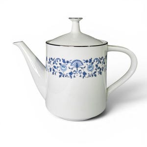 Puede incluir: Una tetera de porcelana blanca con cuerpo cilíndrico y tapa a juego con pomo. La tetera está decorada con una banda horizontal de motivos florales y vegetales en azul, realzada por un fino borde plateado en la parte superior del cuerpo.