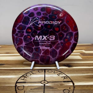 May include: A purple, red and black Prodigy MX-3 midrange disc golf disc with a swirl pattern. The disc is on a white stand and a wooden surface with a burnt-in logo.