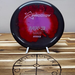 Può includere: Un disco da golf Prodigy MX-3 nero e rosso con una finitura che brilla al buio. Il disco ha un design a spirale rosa e rosso.