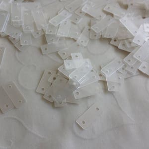 May include: A large quantity of small, white, rectangular sequins. The sequins are scattered on a white surface.