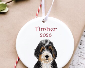 May include: A round white ceramic ornament featuring a watercolor illustration of a tricolor Bernese Mountain Dog puppy. The text Timber and the year 2026 are printed in red above the dog. A thin white ribbon is attached for hanging purposes.
