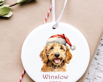 May include: A round white ceramic ornament featuring a watercolor illustration of a golden-brown dog wearing a red and white Santa hat. Below the dog, the text reads Winslow 2026 in red lettering. The ornament hangs from a thin white ribbon.