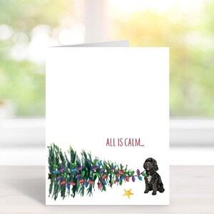 May include: A folded greeting card with a watercolor illustration. The card features a Christmas tree with colorful lights, a black dog, a yellow star, and the text "ALL IS CALM...". The background is a blurred view of a window.