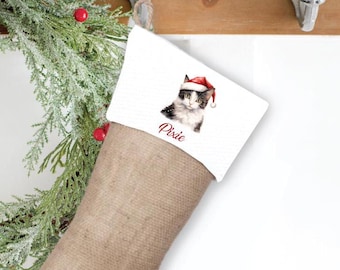 Può includere: Un calzino di Natale personalizzato con uno sfondo bianco che presenta un gatto bianco e nero con un cappello di Babbo Natale rosso. Il nome del gatto, "Pixie", è scritto in corsivo rosso sotto l'immagine.