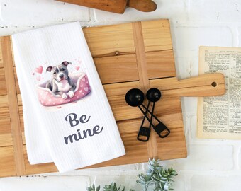 Op de afbeelding: Witte keukendoek met een roze en wit hondenbed met een grijze pitbull puppy erin. De tekst "Be mine" is op de handdoek gedrukt.