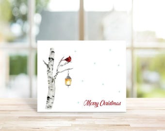 May include: A white greeting card featuring a watercolor illustration of a birch tree branch with a red cardinal bird perched on top and a glowing lantern hanging below. Small blue stars are scattered across the card with Merry Christmas in red.