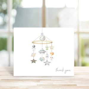 May include: A white card featuring a watercolor illustration of a space-themed mobile. The mobile includes planets, stars, a moon, an astronaut, and a UFO. The words "thank you" are written in script on the card.