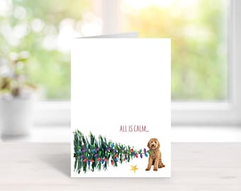 May include: A white greeting card featuring a watercolor illustration of a golden-brown dog sitting next to a horizontal Christmas tree decorated with colorful lights. The text "ALL IS CALM..." is printed in red above the tree and dog.
