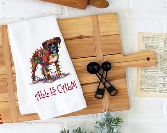 May include: White kitchen towel with a brown and white boxer dog wrapped in Christmas lights. The text "All is Calm" is printed below the dog.