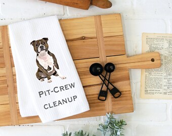 Op de afbeelding: Een witte keukendoek met een bruine en witte pit bull terrier die erop zit. De handdoek heeft de tekst "Pit-Crew Cleanup" in zwart gedrukt.