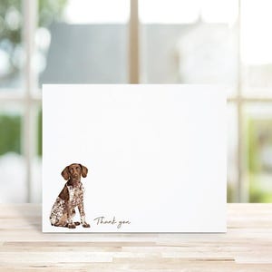 May include: A white thank you card with a brown and white German Shorthaired Pointer dog sitting on the card. The text "Thank you" is printed in brown on the card.