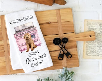 May include: White kitchen towel with a pink oven graphic and a Goldendoodle illustration. Text reads "NO KITCHEN IS COMPLETE WITHOUT A Goldendoodle AT YOUR FEET." The towel is on a wooden cutting board with black measuring spoons.