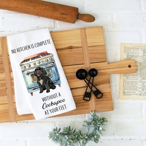 May include: White kitchen towel with a black and white illustration of a dog in front of a blue vintage stove. The text reads "No kitchen is complete without a Cockapoo at your feet."