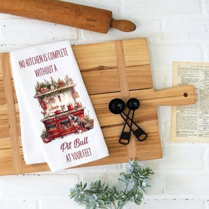 May include: White kitchen towel with a red and white illustration of a vintage kitchen with a dog in front of the stove. The text on the towel reads "No kitchen is complete without a Pit Bull at your feet."