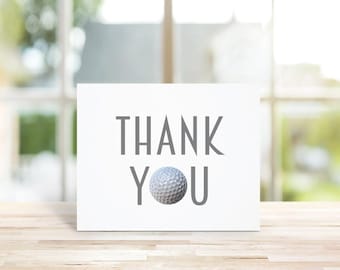 May include: A white card with the words "THANK YOU" in gray text. A golf ball is centered between the words.