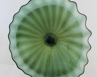 May include: A large, green glass wall sculpture with a wavy, ruffled edge. The glass is textured and has a dark green center.