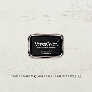May include: A black ink pad with a silver metal casing. The ink pad is labeled "VersaColor" and "Black/Negra/Noire" with the brand name "TSUKINEKO".
