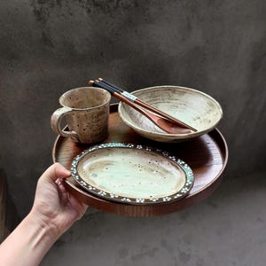 Op de afbeelding: Een houten serveerblad bevat een keramische mok, kom en ovale schaal. Eetstokjes en een houten lepel rusten in de kom. De schaal heeft een bloemenpatroon rond de rand. De mok en kom hebben een gespikkelde, aardse uitstraling.