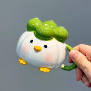 Pode incluir: Uma caneca de cerâmica extravagante em forma de patinho. A caneca é branca com detalhes verdes, pés e bico amarelos e bochechas rosadas. A alça é verde.