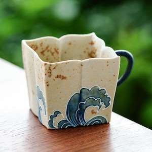 May include: A square ceramic mug with a cream-colored exterior and a dark blue handle. The mug features a blue and white wave design on the front. The mug has a unique, scalloped rim and a rustic, speckled texture.