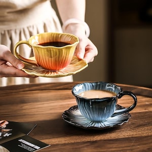 Op de afbeelding: Twee keramische theekopjes en schotels. Eén kopje is geel en oranje met een bijpassende schotel, vastgehouden door een persoon. Het andere kopje is blauw en zwart met een bijpassende schotel. Beide kopjes hebben een bloemenpatroon.