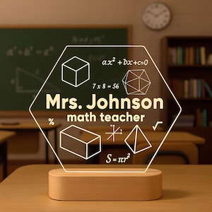 Op de afbeelding: Gepersonaliseerde LED-bureaulamp met een houten voet. De lamp heeft een zeshoekig acryl paneel met de naam "Mrs. Johnson" en "math teacher" en wiskundige symbolen en vergelijkingen. De achtergrond toont een klaslokaal.
