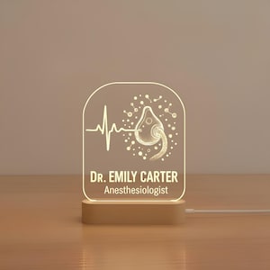 Puede incluir: Lámpara de escritorio personalizada de acrílico con base de madera. La lámpara presenta un tema médico con una línea de latido cardíaco, una mascarilla de oxígeno y el texto "DR. EMILY CARTER Anesthesiologist" en blanco.